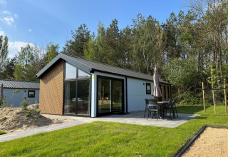 Lodge moderne avec grandes baies vitrées, terrasse avec mobilier et parasol, entouré de pelouse et d’arbres.