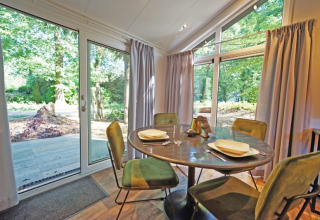 Salle à manger lumineuse dans un lodge avec quatre chaises vertes, table ronde et grandes fenêtres sur la forêt.