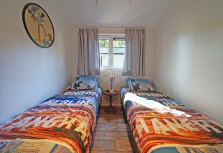 Lichte lodgekamer met twee enkele bedden, kleurrijke dekens, een klein tafeltje en een raam met gordijnen.