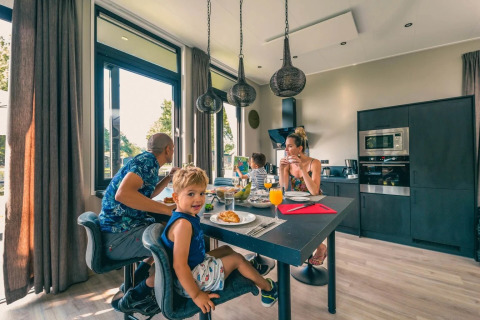 Gezin geniet van ontbijt in moderne glamping accommodatie met grote ramen en zwarte keuken in vakantiepark.