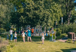 Kinderen en volwassenen genieten van buitenspelen en activiteiten in een zonnig vakantiepark met veel bomen.
