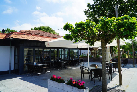 Terras met zitplaatsen, parasols en bomen bij een vakantiepark met glamping, blauwe lucht en veel groen.