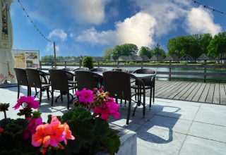 Terras met rieten stoelen en tafels, felgekleurde bloemen, zicht op het water en huisjes op vakantiepark.