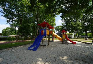 Speeltuin met glijbanen en klimrekken in een vakantiepark met glamping, omringd door bomen en zand.