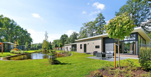 Lodge Grand Modern aan een vijver, omringd door groen gras en bomen op een zonnige dag.