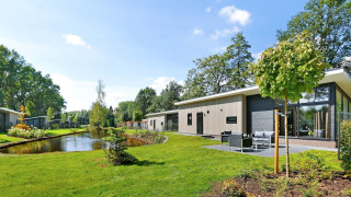 Lodge Grand Modern aan een vijver, te midden van gras en bomen, onder een heldere blauwe lucht.