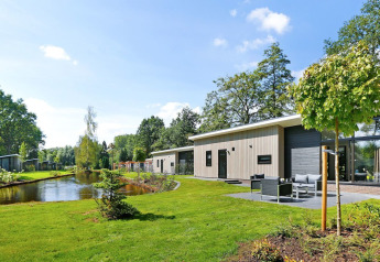 Lodge Grand Modern aan een vijver, omringd door groen gras en bomen op een zonnige dag.