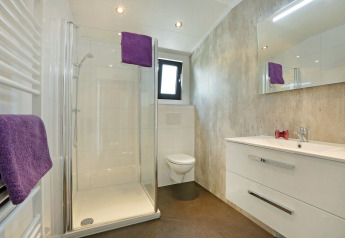 Moderne badkamer met glazen douche, toilet, wit wastafelmeubel en paarse handdoeken in Lodge Grand Modern, De Lochemse Berg.