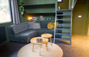 Hébergement de glamping moderne avec canapé vert foncé, tables en bois rondes, tapis et échelle vers mezzanine.