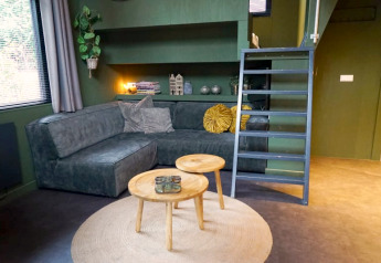 Hébergement de glamping moderne avec canapé vert foncé, tables en bois rondes, tapis et échelle vers mezzanine.