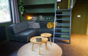 Alojamiento glamping moderno con sofá verde oscuro, mesas redondas, alfombra y escalera hacia el altillo.