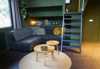 Alloggio glamping moderno con divano verde scuro, tavolini rotondi, tappeto e scala che porta al soppalco.