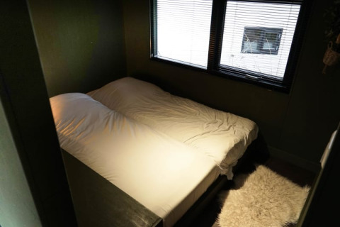 Knusse glamping-accommodatie met tweepersoonsbed, wit beddengoed en grote ramen met jaloezieën.