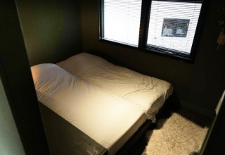 Gemütliches Glamping-Zimmer mit Doppelbett, weißer Bettwäsche und großem Fenster mit Rollläden.