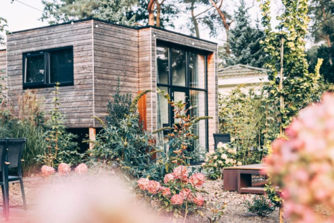 Een modern houten lodgehuis staat tussen bloeiende bloemen en groen in een rustige tuin vol planten.