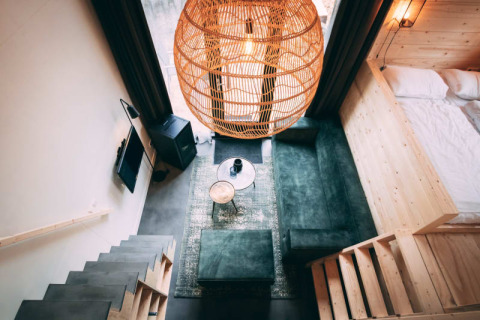 Modern lodge-interieur met groene bank, houten trap, grote rotan lamp en knusse slaaphoek.