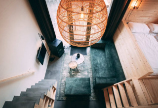 Modern lodge-interieur met groene bank, houten trap, grote rotan lamp en knusse slaaphoek.