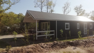 Forest lodge met donkere gevel, witte kozijnen, overdekte veranda en jonge bomen in bosrijke omgeving.