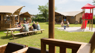 Adultos sentados en mesa de picnic y niños jugando en parque frente a tiendas safari en Camping 't Veld.