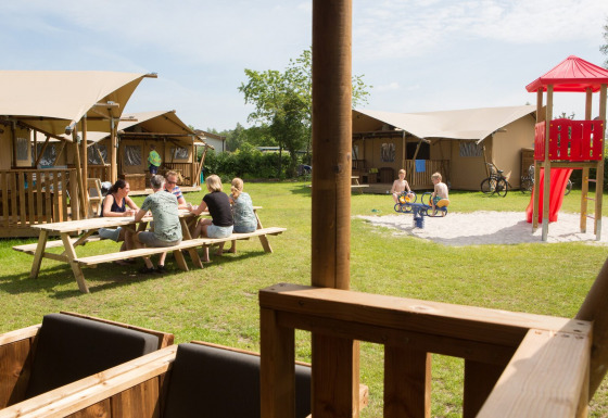 Volwassenen aan een picknicktafel, kinderen spelen op speeltuin vlak bij safaritenten op Camping 't Veld.
