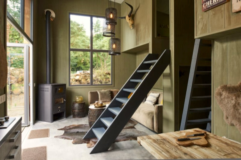 Cozy lodge interior with wood furniture, loft ladders, a wood stove, large windows, and rustic decor.