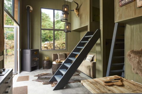 Knusse lodge met houten meubels, ladders naar slaaplofts, houtkachel, grote ramen en rustieke sfeer.