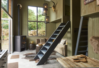 Knusse lodge met houten meubels, ladders naar slaaplofts, houtkachel, grote ramen en rustieke sfeer.