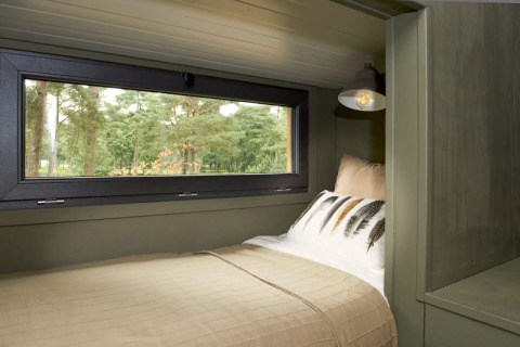 Cozy glamping bed with large window view of the forest, beige bedding, and a modern reading lamp.
