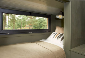 Lit confortable en hébergement glamping avec grande fenêtre sur la forêt, linge beige et lampe de lecture.