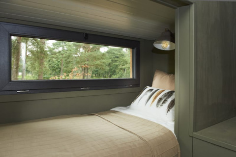 Cama de glamping junto a una gran ventana con vistas al bosque, ropa de cama beige y lámpara moderna.