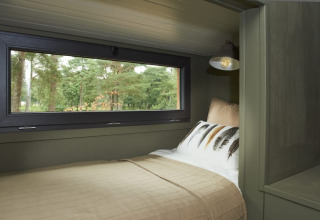 Knus glampingbed met groot raam met uitzicht op het bos, beige beddengoed en moderne leeslamp.