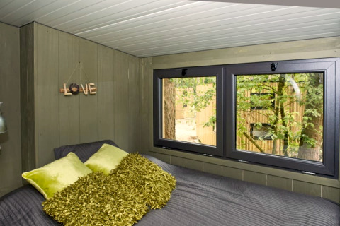 Cozy lodge bedroom with green pillows and textured blanket, large windows overlooking trees outside.