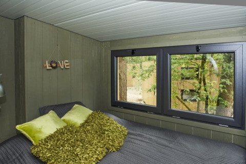 Knusse lodgeslaapkamer met groene kussens, grote ramen en uitzicht op bomen buiten.