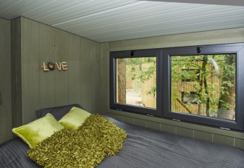 Dormitorio acogedor de lodge con cojines verdes y manta, ventanales con vista a árboles exteriores.
