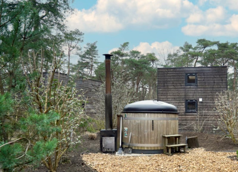 Outdoor wooden hot tub and cabins surrounded by woodland at a lodge, beneath a blue sky with clouds.