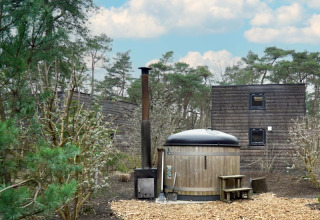 Outdoor wooden hot tub and cabins surrounded by woodland at a lodge, beneath a blue sky with clouds.