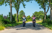 To personer kører el-scootere gennem en grøn allé på en solrig dag i en feriepark med glamping.