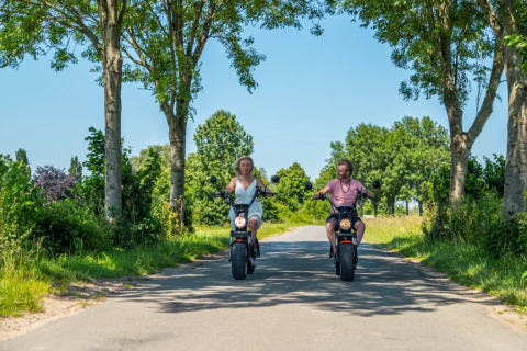To personer kører el-scootere gennem en grøn allé på en solrig dag i en feriepark med glamping.