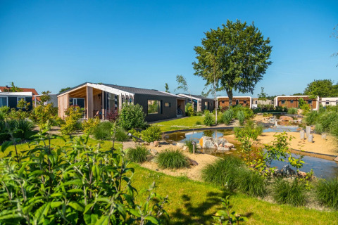 Imagen de un parque vacacional con alojamientos glamping, abundante vegetación y estanque bajo cielo azul.
