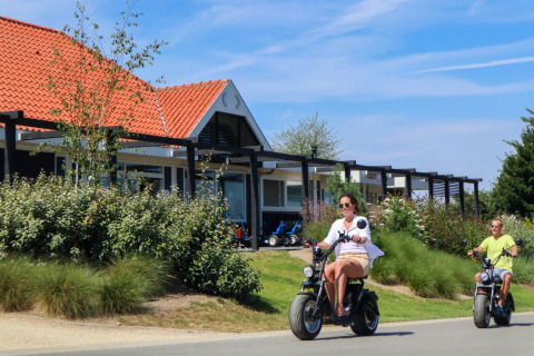 To personer kører på el-scootere forbi glamping-hytter i en feriepark med røde tegltage og grøn beplantning.