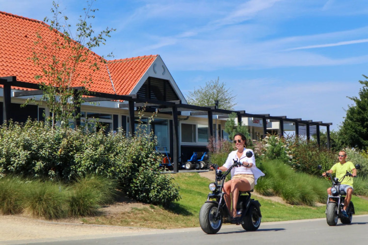 Two people ride electric scooters past glamping cabins at a holiday park with red tiled roofs and greenery.