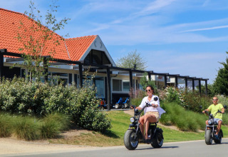 Two people ride electric scooters past glamping cabins at a holiday park with red tiled roofs and greenery.