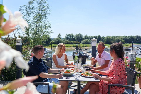 Cuatro personas disfrutan de una comida al aire libre en una terraza junto a un puerto deportivo en un parque de glamping.