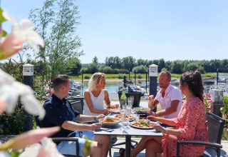 Fire mennesker nyder et måltid udendørs på en terrasse ved en havn i en feriepark med glamping.