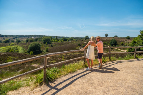 Un couple admire la vue près d’une clôture dans un parc de vacances avec glamping sous un ciel ensoleillé.