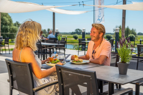Koppel luncht buiten op een terras in een vakantiepark met glampingaccommodaties aan het water.