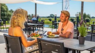 Una pareja disfruta de un almuerzo al aire libre en un parque vacacional con alojamientos de glamping junto al río.