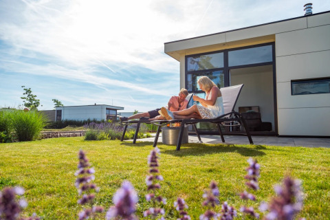 Paar entspannt auf Sonnenliegen vor einer modernen Glamping-Unterkunft im Ferienpark mit Garten.