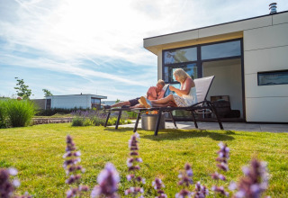 Par nyder solen på liggestole foran moderne glampingbolig i en feriepark med velplejet græsplæne.