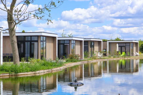 Cabañas modernas de glamping junto a un estanque en un parque vacacional rodeado de naturaleza y cielo azul.